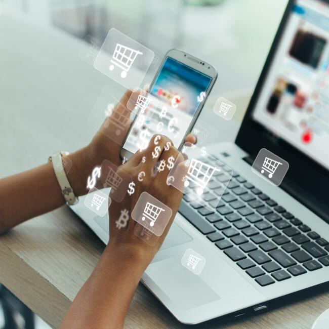 A person online shopping on their phone and laptop. Photo is overlaid with dollar signs and shopping cart icons. 