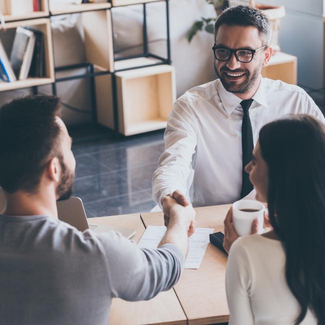 A banker sits across from two clients, shaking hands with one of them. 