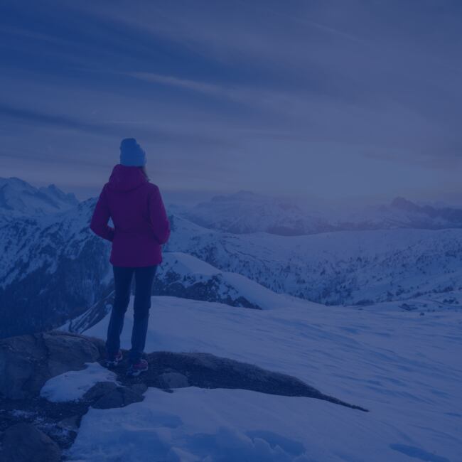 A person stands on a hilly landscape overlooking snowy mountains. 