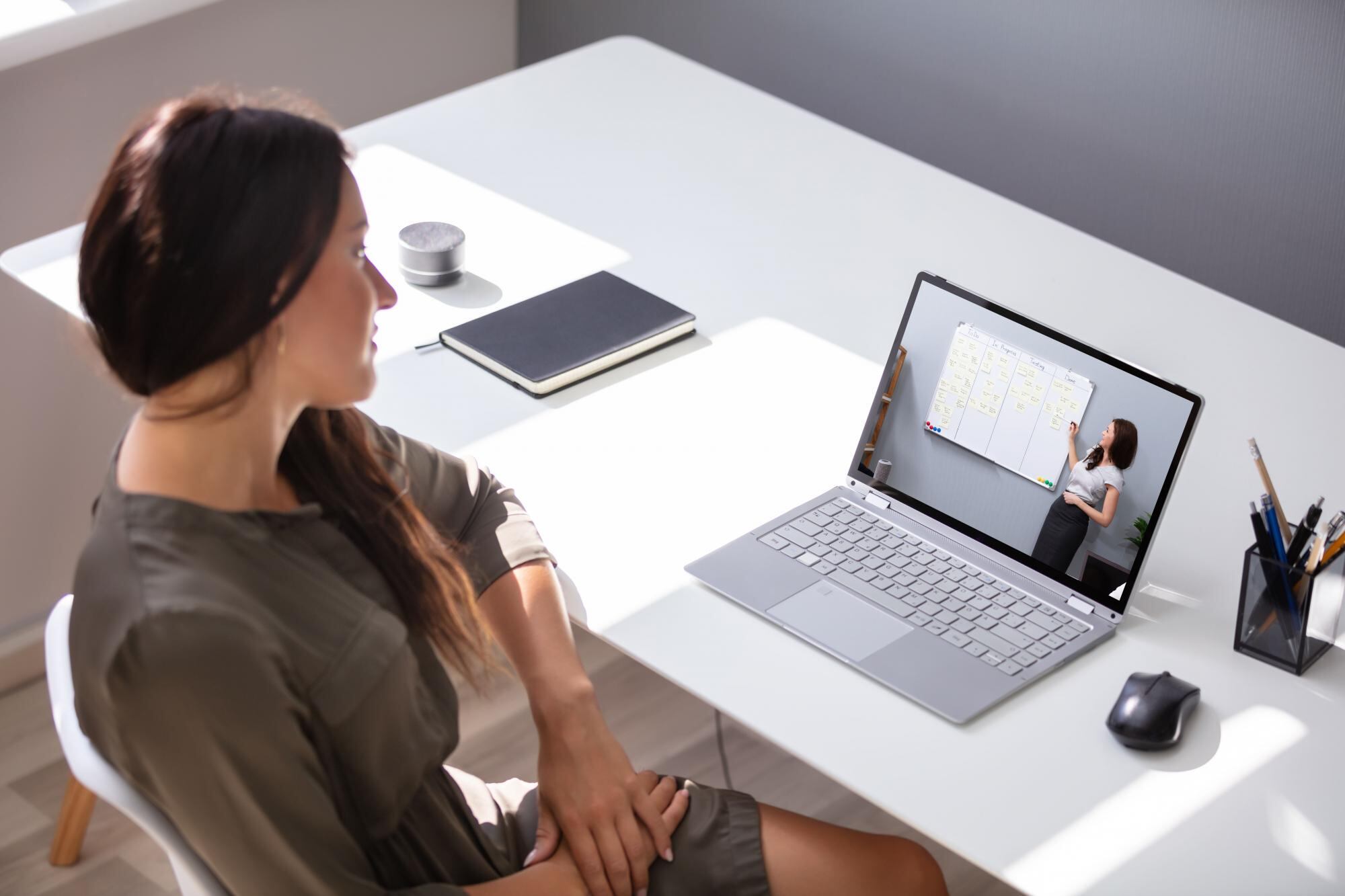 A woman sits at a desk and watches a video on her laptop of a lecturer pointing at a whiteboard. 